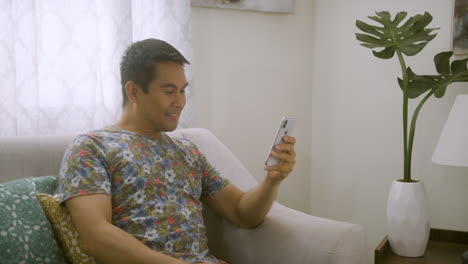 Asian-Man-Smiling-While-Browsing-On-His-Phone,-Sitting-On-A-Couch