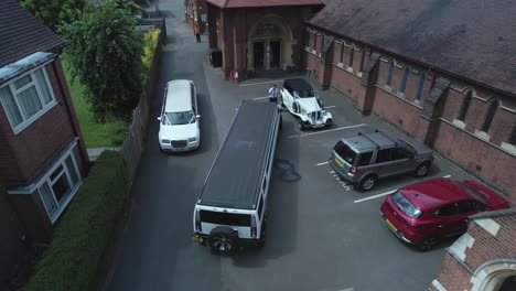 Expensive-wedding-limousine-vehicles-parked-outside-charming-English-church-aerial-descending-view