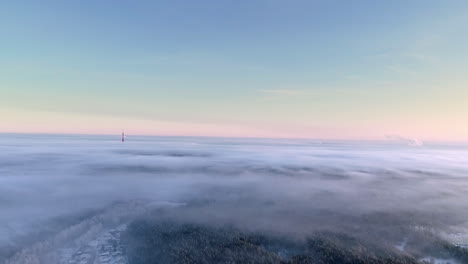 Beautiful-aerial-backward-movement-shot-over-a-fog-covering-a-forest-with-the-view-of-a-tv-tower-in-the-distance-in-the-morning-sunlight