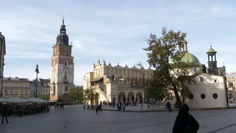 Gente-Paseando-En-La-Plaza-Del-Mercado-Principal-De-Cracovia-En-Polonia-Con-Vistas-A-La-Torre-Del-Ayuntamiento,-A-La-Sala-De-Telas-Y-A-La-Iglesia-De-San