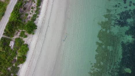 Rising-above-a-solo-traditional-fishing-boat-moored-on-a-white-sandy-beach-shoreline-with-crystal-clear-ocean-water-on-idyllic,-remote-tropical-island-paradise