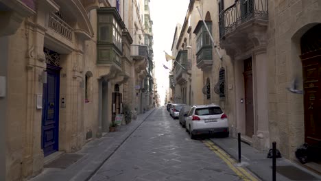 Coches-Aparcados-En-Una-Calle-Estrecha-En-La-Ciudad-De-Valletta-Con-Banderas-En-Los-Balcones-Ondeando-En-El-Viento