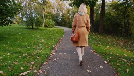 Eine-Frau-In-Einem-Mantel-Mit-Einer-Tasche-Geht-In-Einem-Herbstpark-Spazieren,-Rückansicht