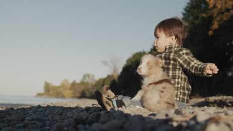 The-child-sits-on-the-shore-of-the-lake,-throws-pebbles-into-the-water.-Next-to-him-is-a-small-puppy.