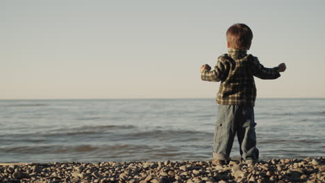 Ein-Zielstrebiger-Zweijähriger-Junge-Wirft-Einen-Stein-Ins-Meer.-Rückansicht,-Zeitlupenvideo