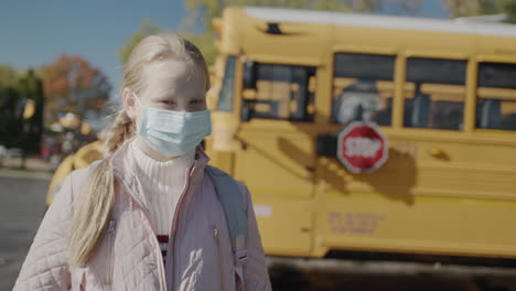 Child-in-a-protective-mask-stands-in-front-of-a-school-bus.-A-Stop-sign-is-visible-at-the-rear.