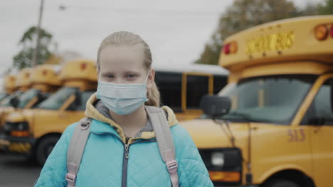Portrait-of-schoolgirl-in-a-protective-mask-against-the-background-of-a-row-of-yellow-buses
