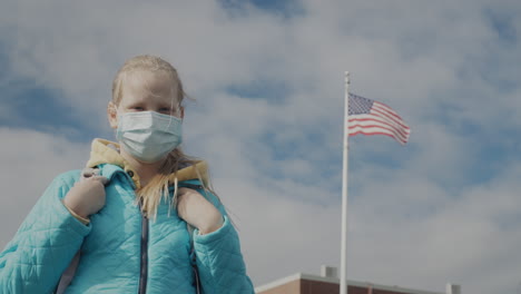 A-child-in-a-protective-mask-stands-against-the-background-of-a-school-and-a-US-flag.