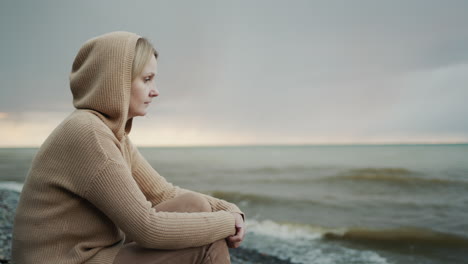A-woman-in-a-warm-sweater-with-a-hood-on-her-head-sits-on-the-ocean-shore,-where-the-dramatic-sky-and-surf.-Side-view