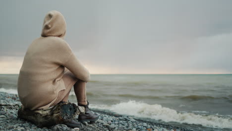 A-woman-in-a-warm-sweater-with-a-hood-on-her-head-sits-on-the-ocean-shore,-where-the-dramatic-sky-and-surf