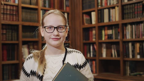 Portrait-of-a-11-year-old-girl-with-a-textbook,-stands-against-the-background-of-bookshelves-in-the-library