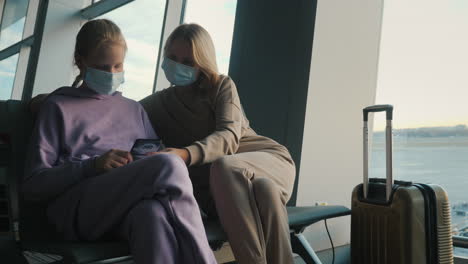 Mother-and-daughter-waiting-for-their-flight-in-the-airport-terminal.-They-are-wearing-protective-masks,-using-a-smartphone