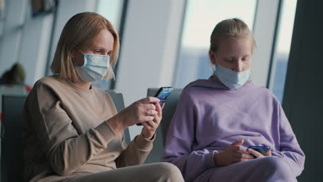 A-parent-with-a-child-is-waiting-for-a-flight-in-the-airport-terminal.-On-the-faces-of-protective-masks,-use-smartphones