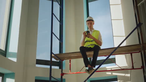 A-worker-uses-a-smartphone,-sits-high-on-scaffolding-inside-the-house