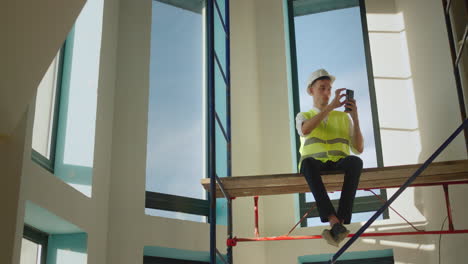A-worker-uses-a-smartphone,-sits-high-on-scaffolding-inside-the-house