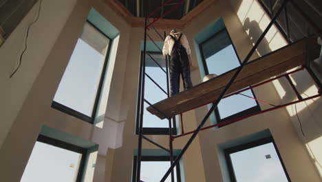 Painter-primes-the-wall,-stands-high-on-the-scaffolding-inside-the-house.-Low-angle-shot
