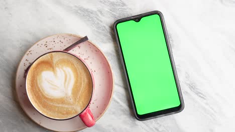 Smart-phone-with-green-screen-and-cup-of-coffee-on-table-,