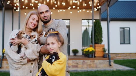 Portrait-of-a-happy-family-against-the-backdrop-of-their-home.-Mom,-dad-and-daughter---holding-several-little-beagle-puppies-in-their-hands
