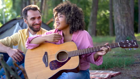 Happy-hiker-couple-camping-in-the-forest.-Young-black-woman-plays-guitar-and-sings,-caucasian-man-smiles.