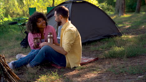 Happy-couple-have-a-hot-coffee-with-a-thermos-and-a-metal-mug-camping-in-the-forest.-Caucasian-man-and-young-black-female-outdoors.