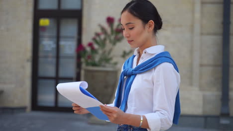 Asian-businesswoman-working-on-the-street-reading-documents-on-a-clipboard.-Medium-shot.-Copy-space.-Moving-camera.