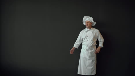 Male-chef-with-hand-aside,-showing-an-empty-space-on-his-palm-for-advertising-or-commercial-text,-smiling-and-looking-at-camera.-Copy-space-and-black-background.