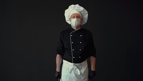 Male-chef-ready-to-cook,-wearing-medical-face-mask-and-gloves,-showing-cooking-tools,-frying-pan-and-slotted-turner.-Medium-shot-on-black-background.