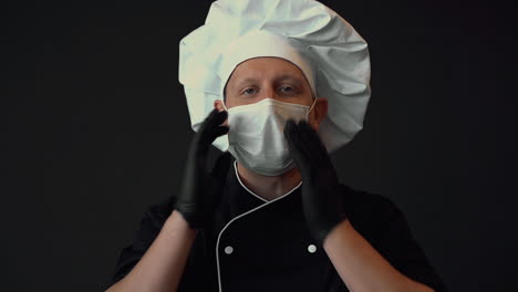 Male-chef-ready-to-cook,-puts-on-medical-face-mask-and-and-makes-the-OK-gesture.-Portrait-on-black-background.