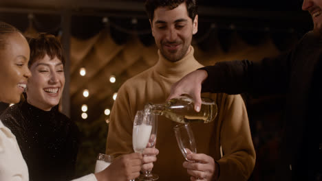 Happy-caucasian-man-pouring-champagne-in-his-cheerful-friends'-glasses-at-New-Year's-Eve-party