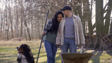 Caucasian-couple-and-their-dog-holding-a-wheelbarrow-and-a-rake-in-the-countryside