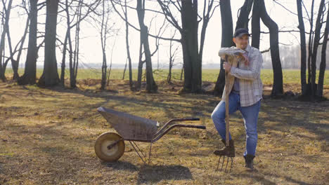 Caucasian-man-leaning-on-the-stick-of-a-rake-standing-near-a-wheelbarrow-in-the-countryside