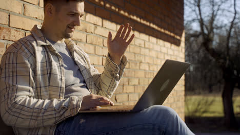 Close-up-view-of-caucasian-man-having-a-video-call-on-laptop-while-sitting-on-a-bench-outside-a-country-house