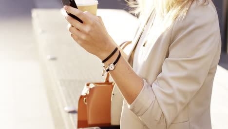 Attractive-blonde-business-woman-using-smartphone-drinking-coffee-commuter-in-city-london