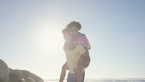 Attractive-man-giving-girlfriend-piggyback-mixed-race-couple-enjoying-nature-on-the-beach