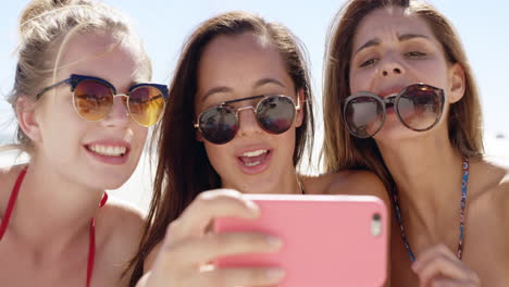 Three-teenage-girl-friends-taking-selfie-on-beach-blowing-kiss-pulling-funny-faces-sharing-vacation-photo