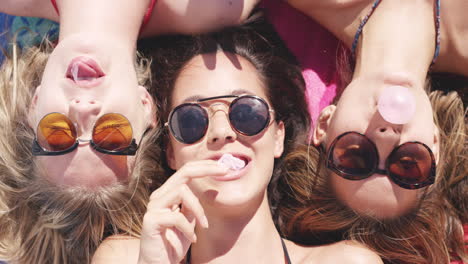 Top-view-of-three-teeanage-girl-friends-lying-on-back-blowing-bubblegum-candy-bubbles-on-beach