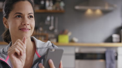 Pretty-mixed-race-woman-using-touch-screen-smart-phone-at-home-in-kitchen-wearing-sportswear-drinking-coffee-smiling