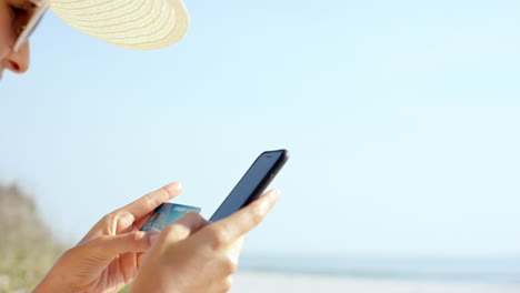Woman-using-credit-card-shopping-online-with-mobile-phone-at-the-beach-on-vacation-over-the-shoulder-shot