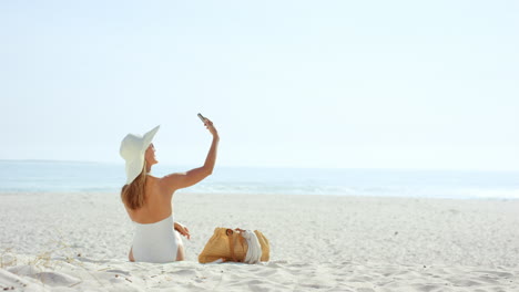Woman-talking-selfie-using-phone-sitting-on-beach-wearing-designer-one-piece-swimsuit