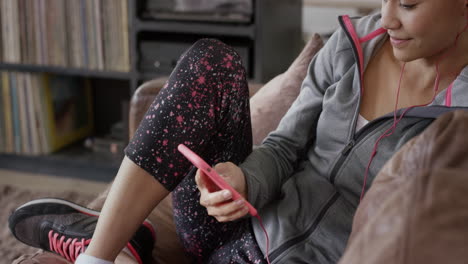 Sporty-woman-listening-to-music-browsing-internet-on-smart-phone-at-home-relaxing-on-couch