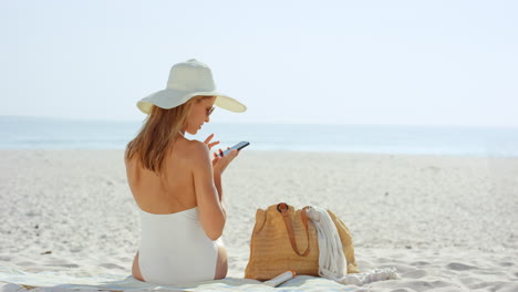 Woman-talking-selfie-using-phone-sitting-on-beach-wearing-designer-one-piece-swimsuit