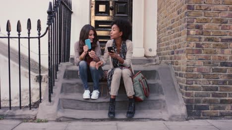 Two-beautiful-woman-friends-sitting-on-steps-having-fun-using-smart-phone
