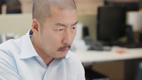 Japanese-Businessman-executive-working-at-computer-using-digital-tablet-connected-to-global-data-in-busy-corporate-office-committed-to-growth-and-success