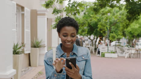 Retrato-De-Una-Elegante-Mujer-Afroamericana-Sonriendo-Feliz-Disfrutando-De-Mensajes-De-Texto-Navegando-En-Línea-Usando-Un-Teléfono-Inteligente-En-La-Ciudad