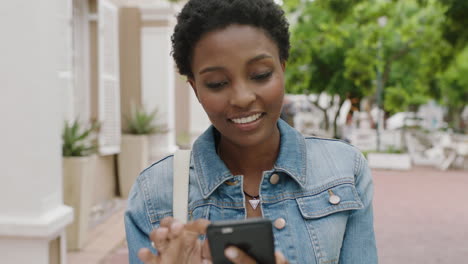 Retrato-De-Una-Elegante-Mujer-Afroamericana-Sonriendo-Disfrutando-De-Mensajes-De-Texto-Navegando-En-Línea-Usando-Un-Teléfono-Inteligente-En-La-Ciudad