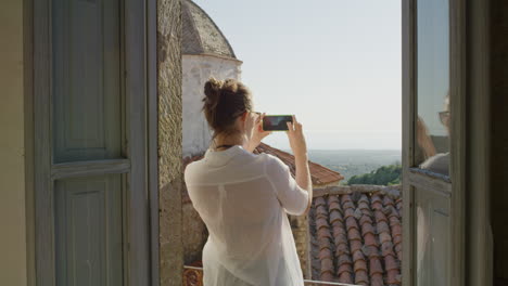 Glückliche-Frau,-Die-Ihr-Smartphone-Nutzt,-Um-Ein-Foto-Zu-Machen-Und-Es-Zu-Genießen,-Sommerurlaubsreiseerlebnisse-Zu-Teilen-Und-Die-Wunderschöne-Aussicht-Auf-Dem-Balkon-Mit-Sonnenbrille-Zu-Fotografieren