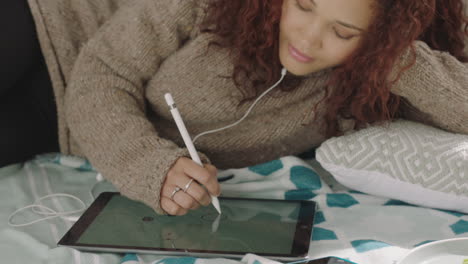 young-mixed-race-woman-designer-using-tablet-pc-stylus-drawing-on-touchscreen-sketching-creative-ideas-relaxing-on-bed-listening-to-music