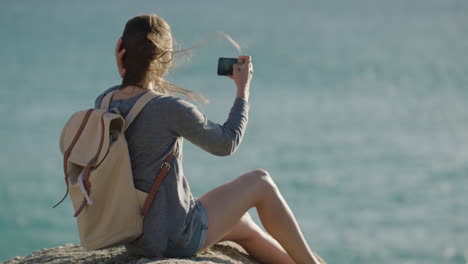 Attraktive-Junge-Frau-In-Zeitlupe,-Die-Mithilfe-Der-Smartphone-Kameratechnologie-Ein-Foto-Des-Ruhigen-Meeres-Am-Meer-Macht,-Sitzt-Entspannt-Und-Genießt-Den-Sommerwind,-Der-Durch-Die-Haare-Weht