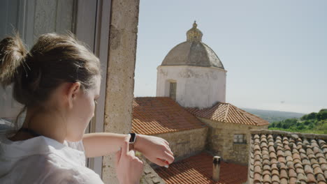 Mujer-Usando-Un-Reloj-Inteligente-Revisando-Mensajes-Disfrutando-De-Un-Cálido-Día-Soleado-De-Vacaciones-De-Pie-En-El-Balcón-Turista-Feliz-Relajándose-En-Italia