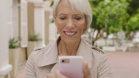 Primer-Plano-Retrato-De-Una-Anciana-Elegante-Sonriendo-Feliz-Disfrutando-De-Mensajes-De-Texto-Navegando-Usando-Un-Teléfono-Inteligente-En-La-Ciudad